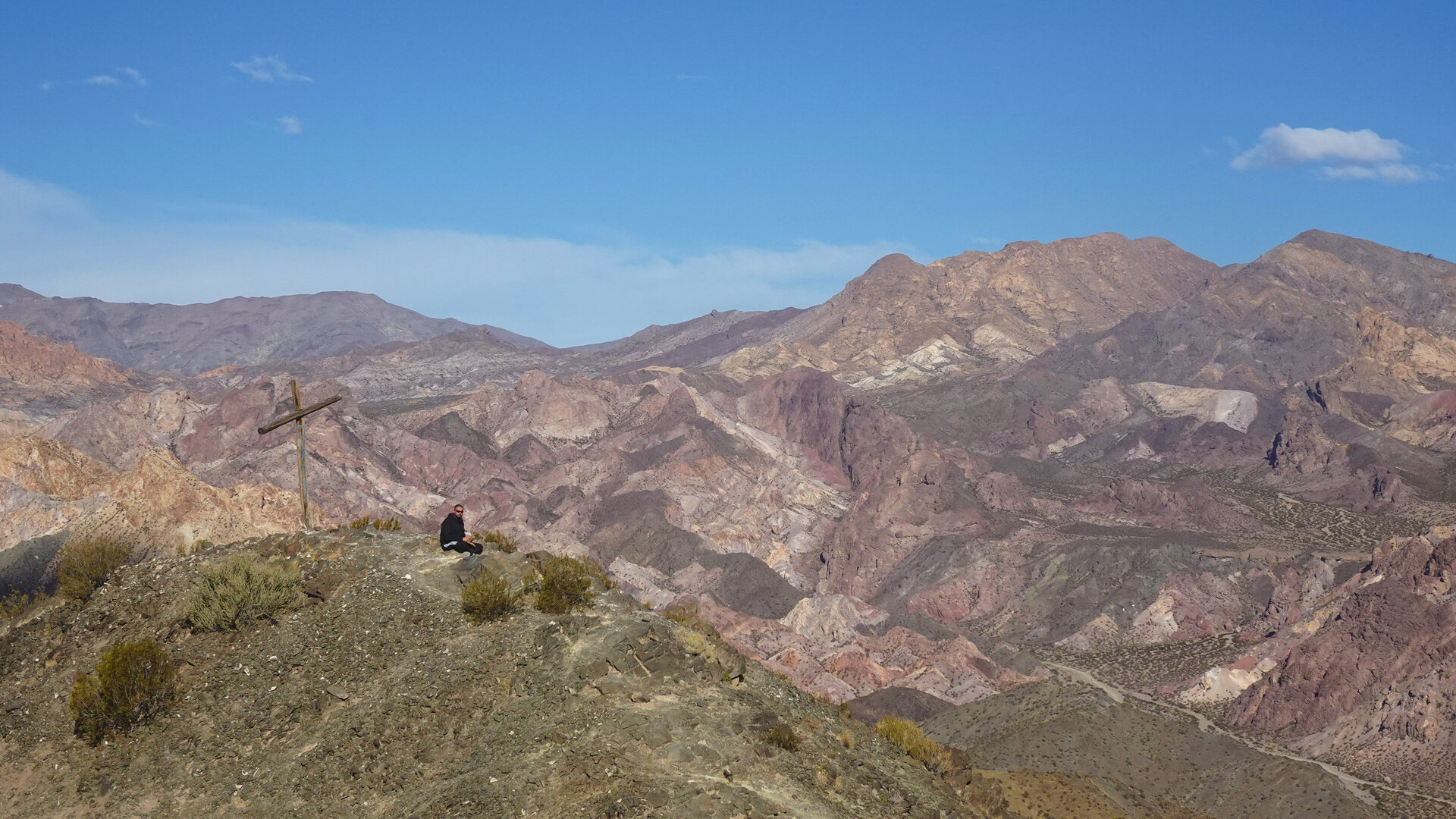 A person sitting beside a wooden cross above folded red desert mountains