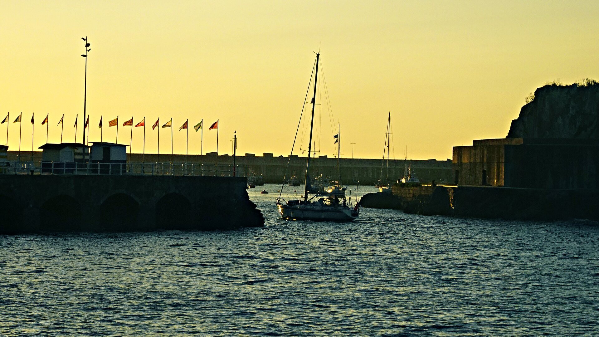 A yacht leaving a flag-lined harbour mouth at sunset