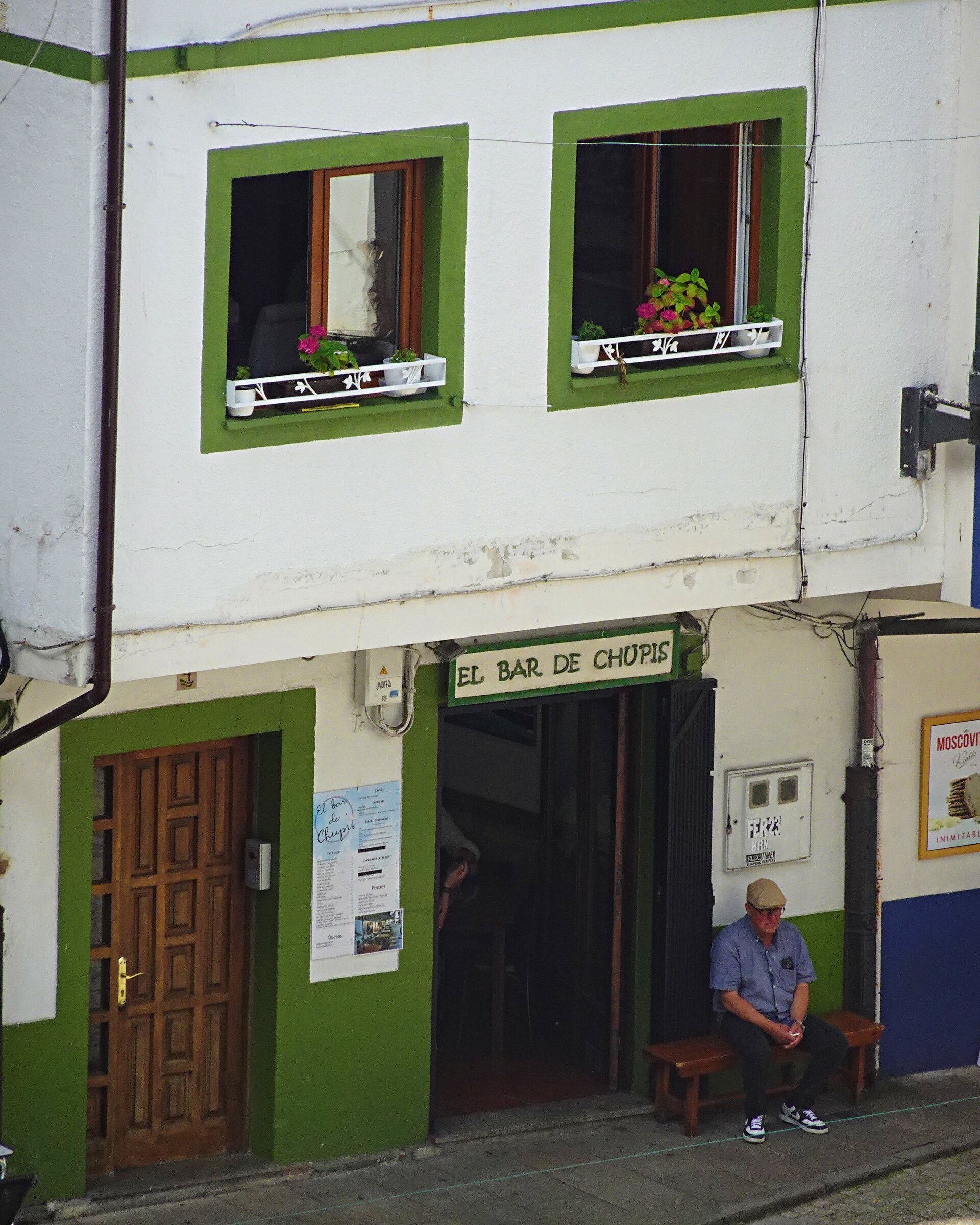 A man sitting on a bench outside a green and white village bar