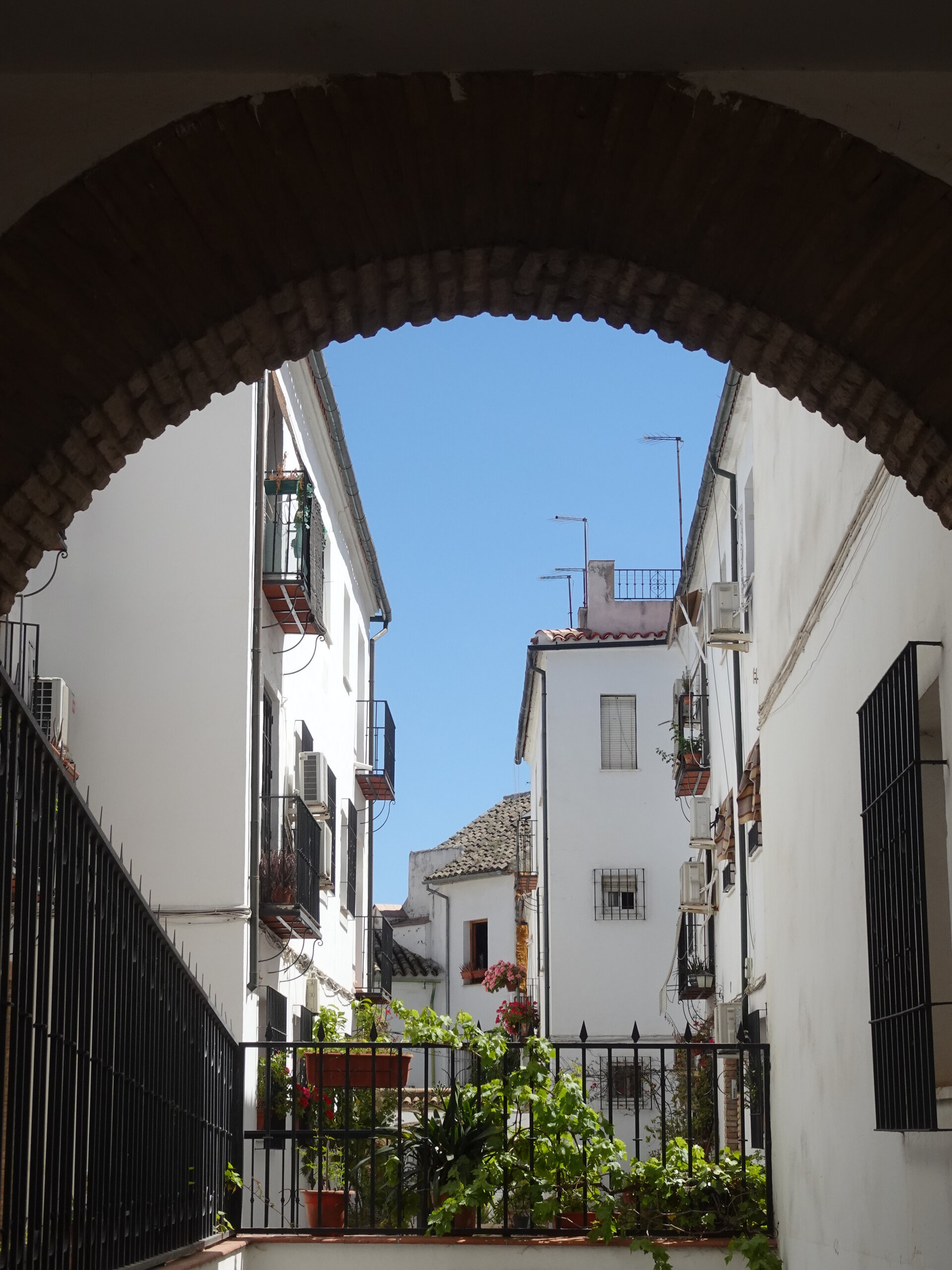 View through a brick archway into a sunlit alley of white houses with iron balconies