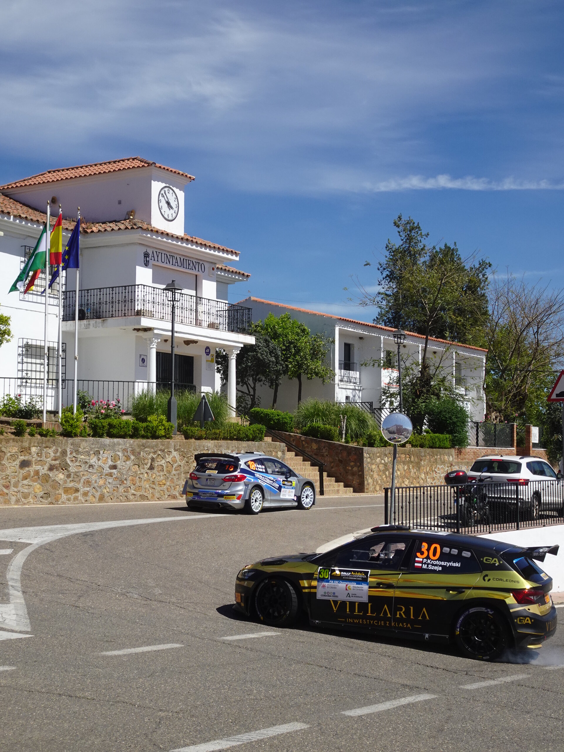 Two rally cars rounding a corner in front of a small whitewashed town hall