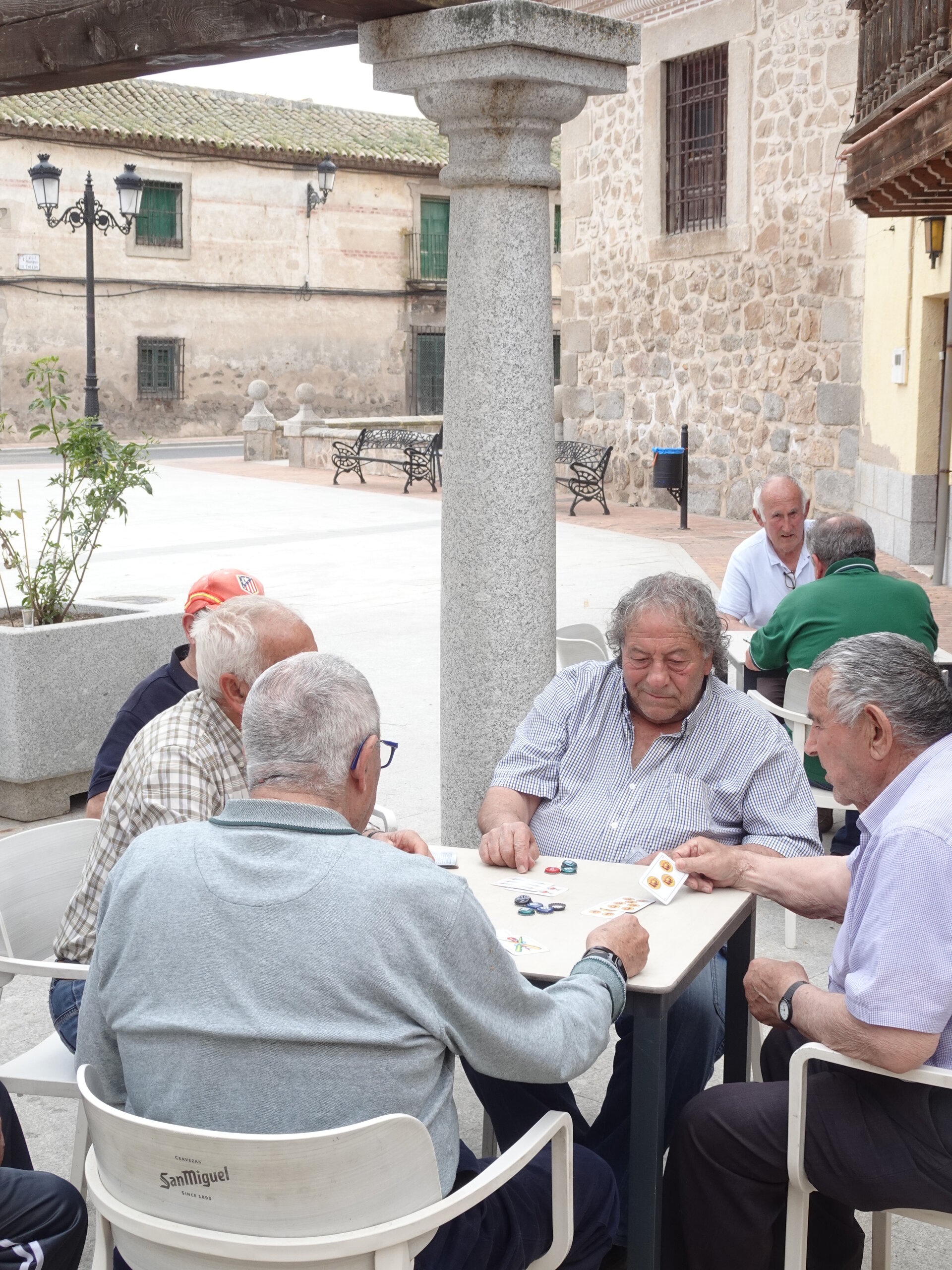Group of older men playing cards at an outdoor cafe table beside a granite column