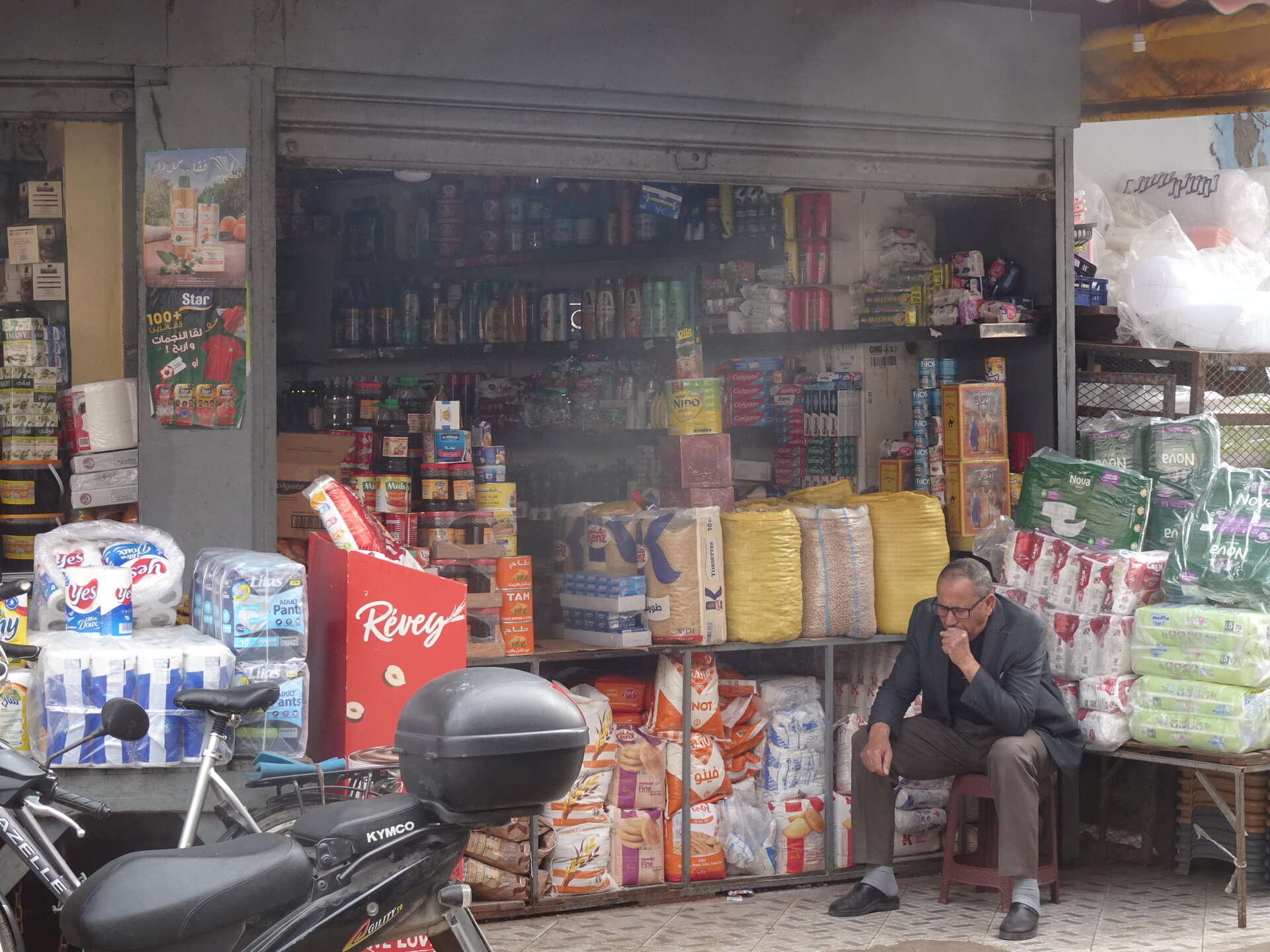 Man sitting outside a densely stocked corner shop with a motorbike parked in front
