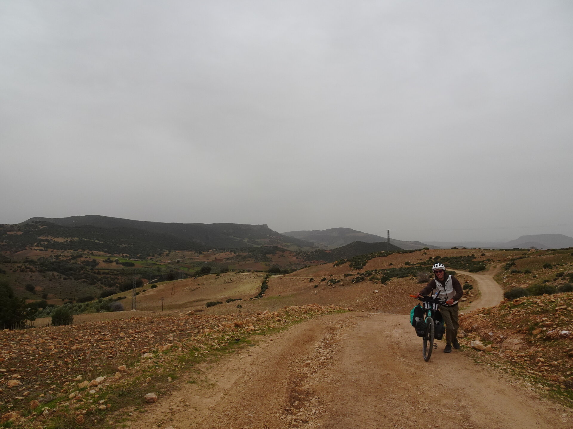 Cyclist pushing a loaded bike up a dirt track through barren hills in fog
