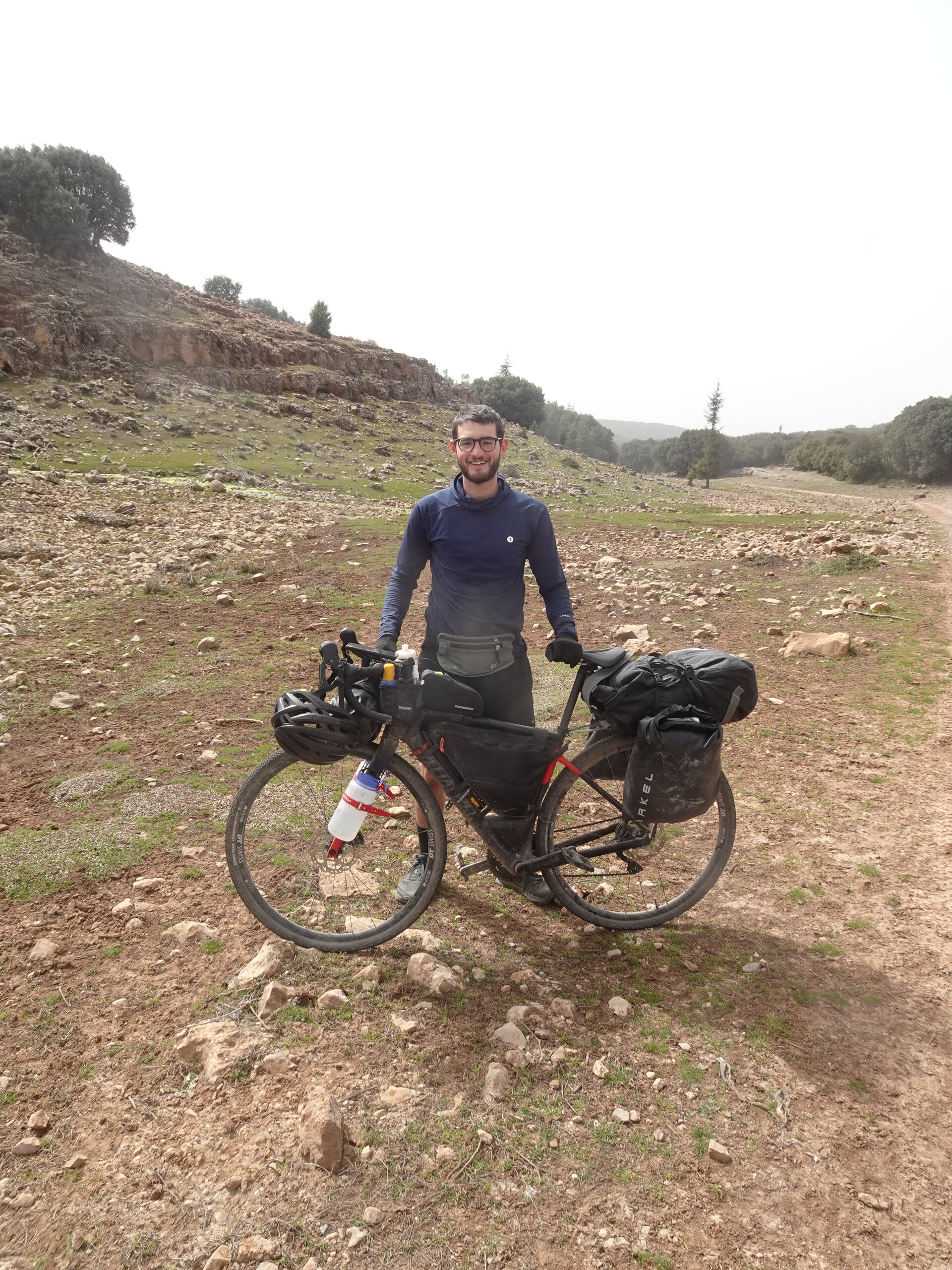 Smiling cyclist posing with loaded bike on a rocky hilltop trail