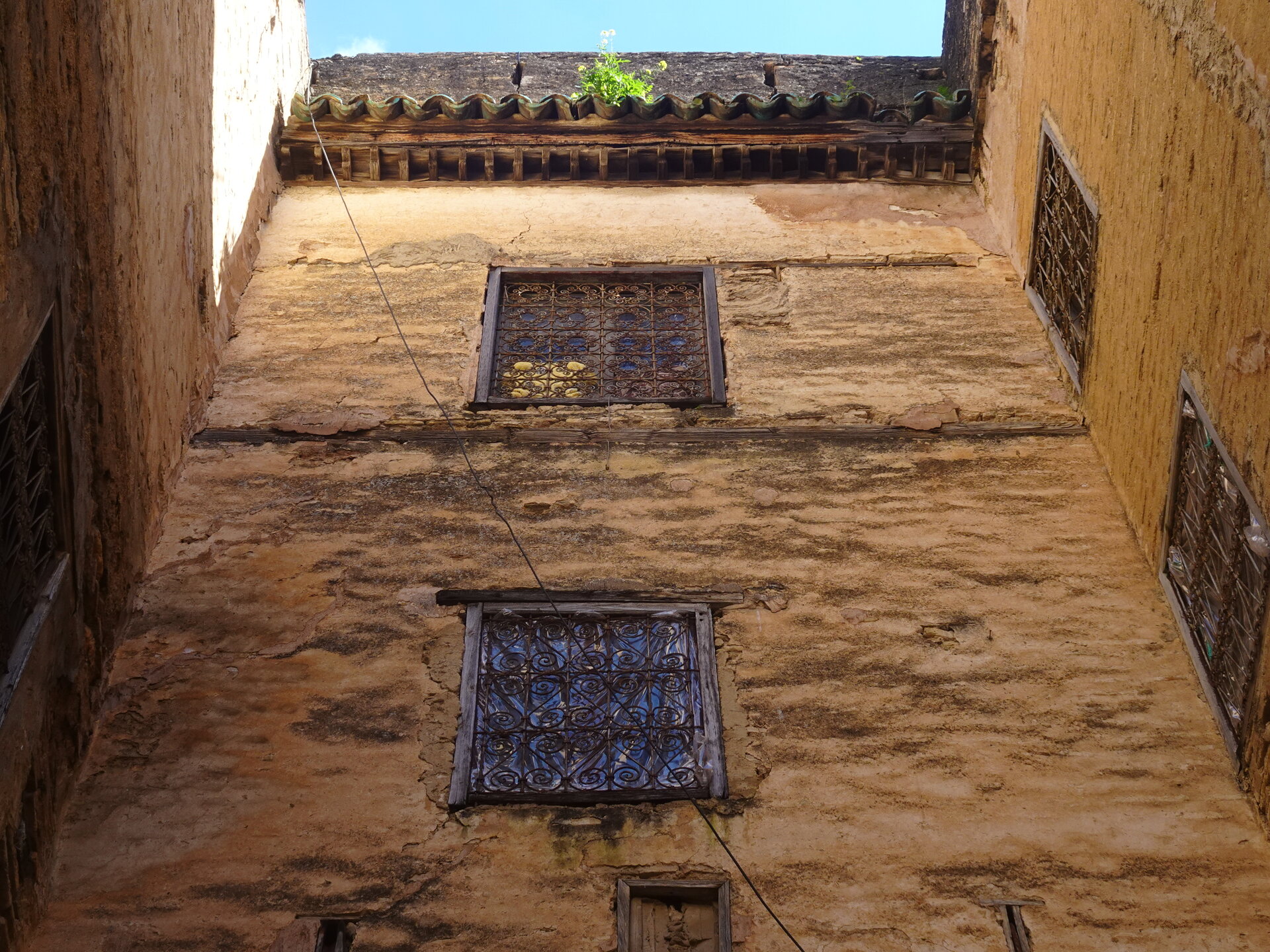 Looking up at weathered mud walls with iron-grille windows in a narrow alley