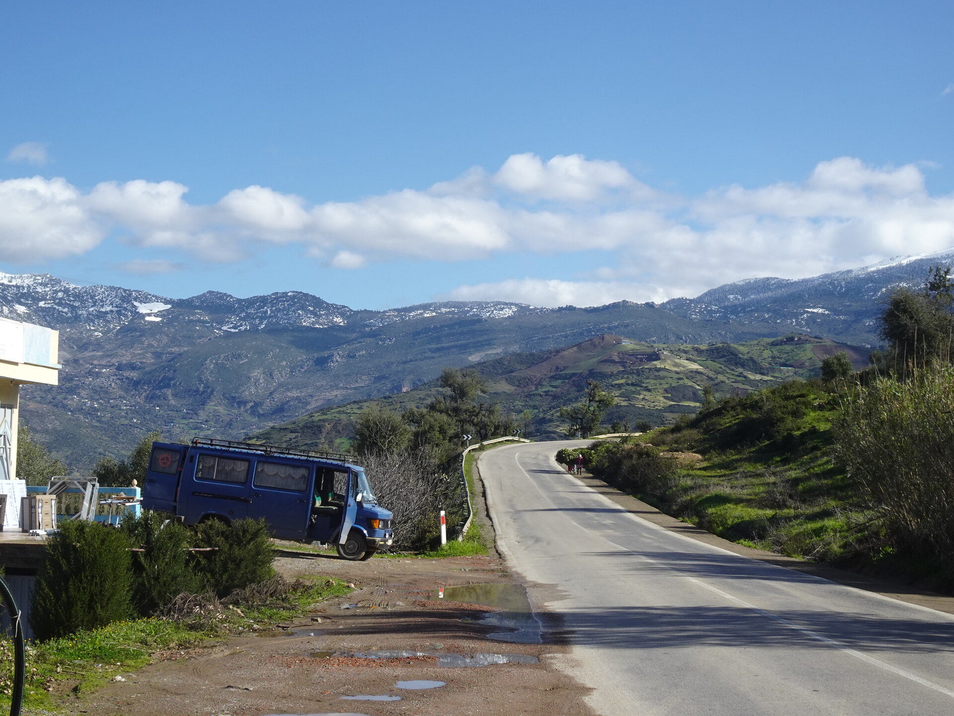 Blue van beside a mountain road winding toward snowy peaks