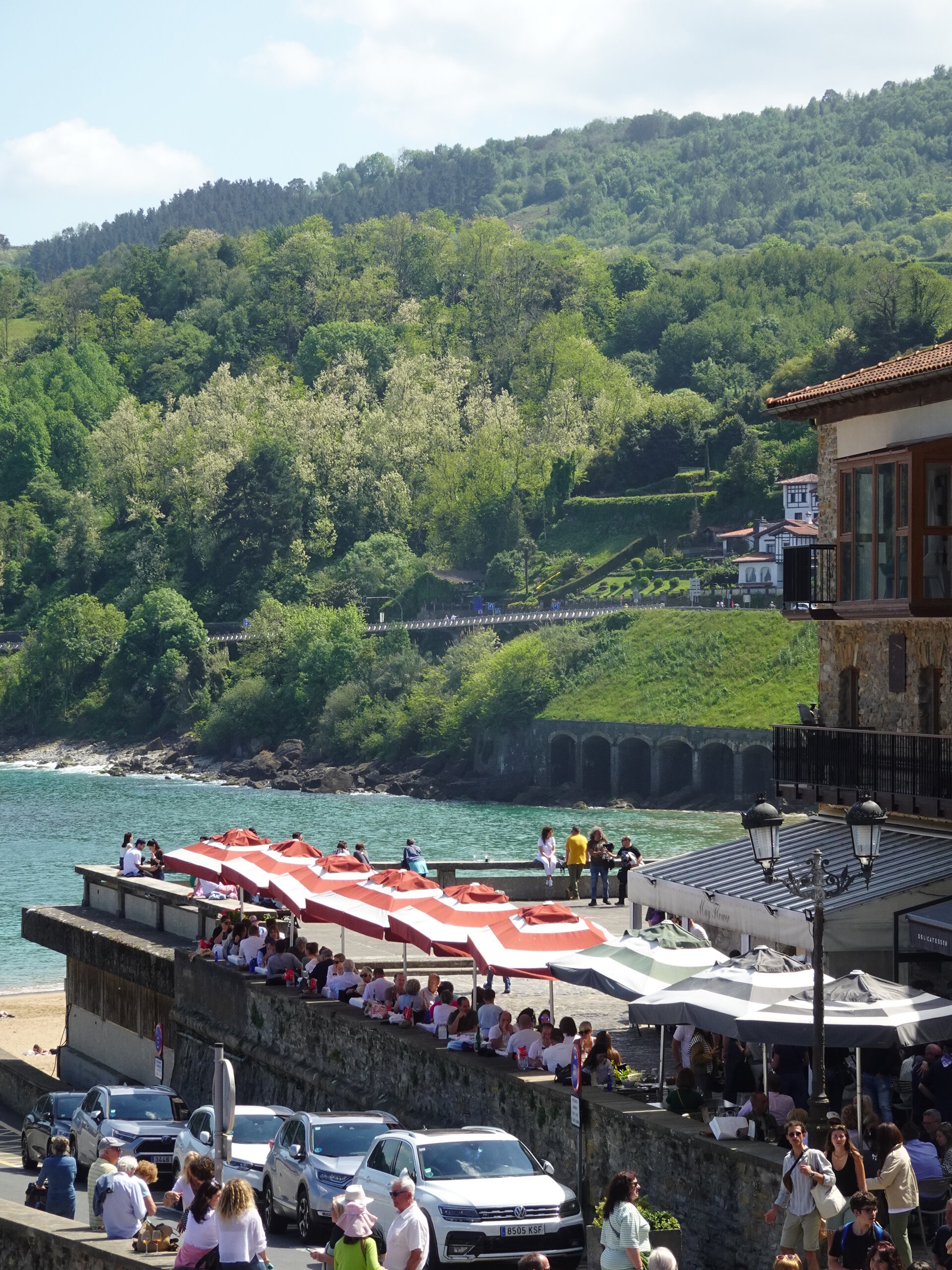 Crowds and striped umbrellas on a seafront below green wooded hills