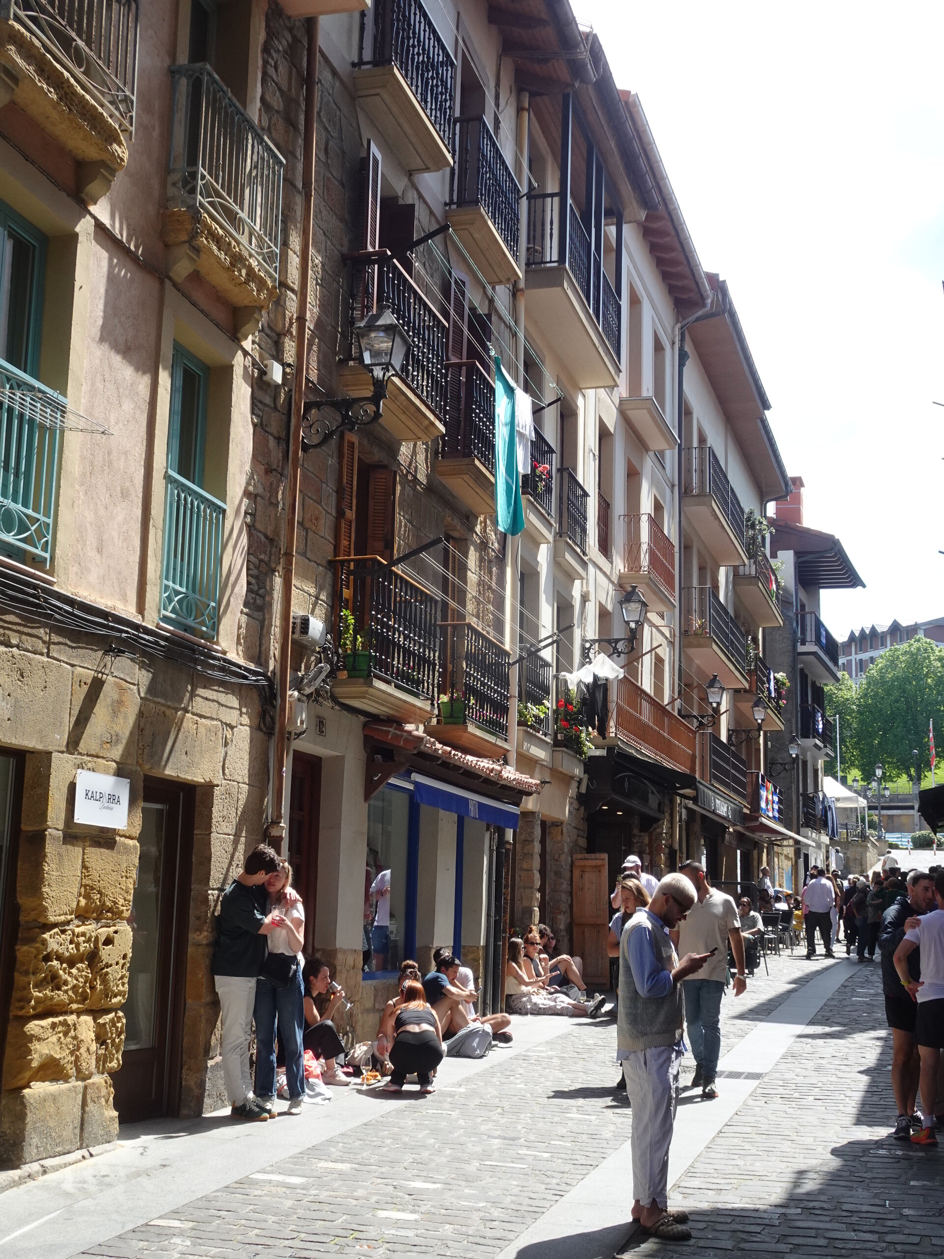 People sitting along a sunny cobbled old-town street with balconied houses