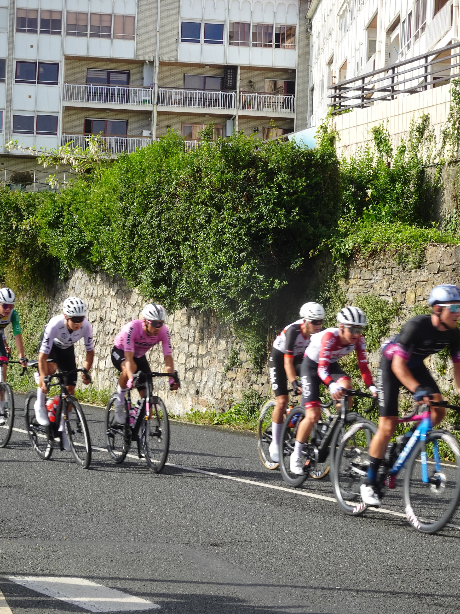 Road racers riding past a stone wall and apartment blocks on a town climb