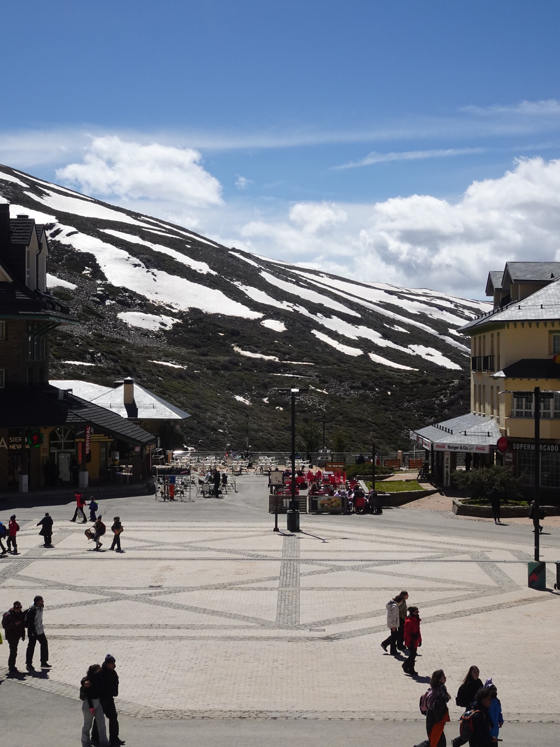 Ski resort plaza at the foot of patchy snow covered slopes under blue sky