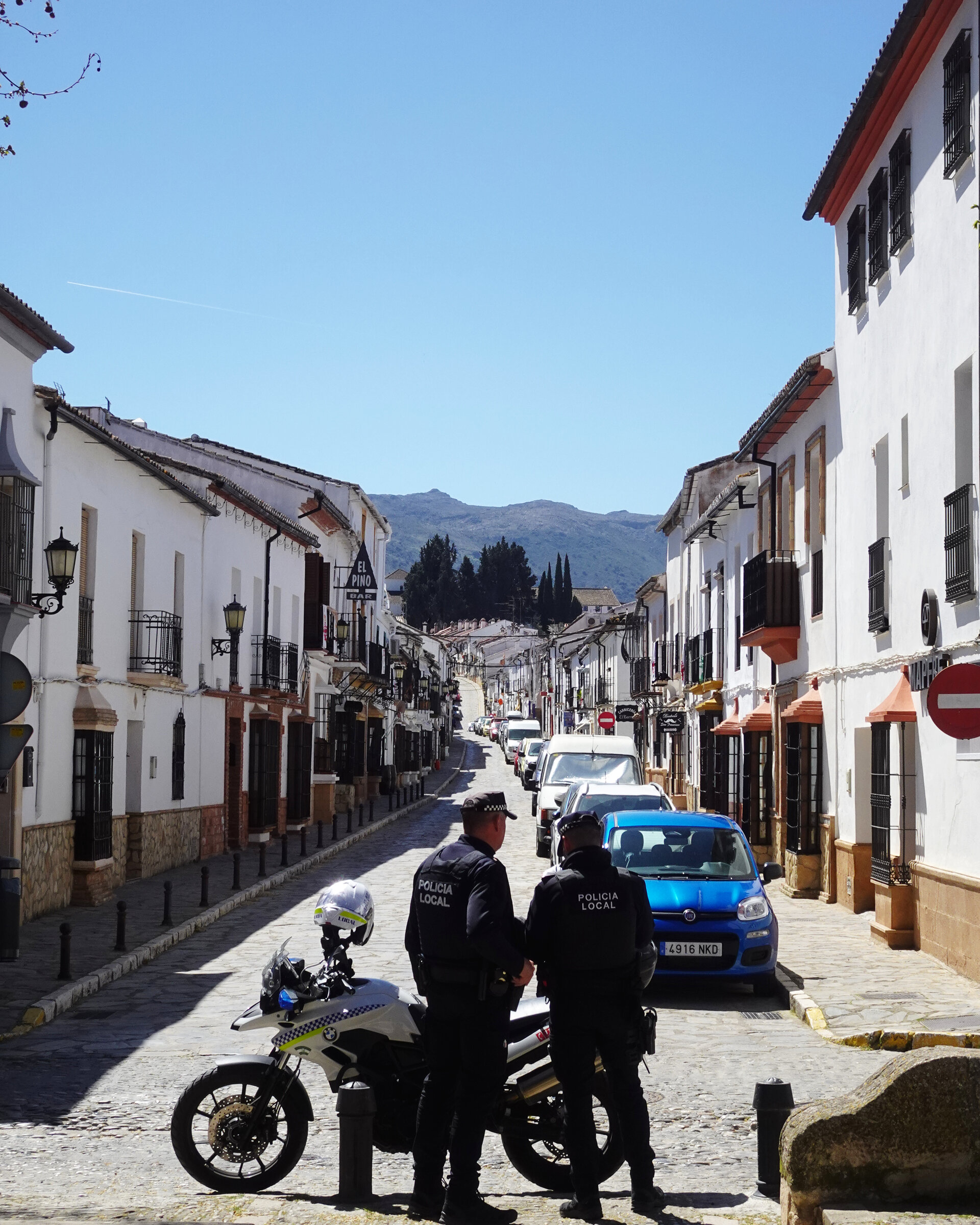A sunlit whitewashed street with a parked scooter and passers-by