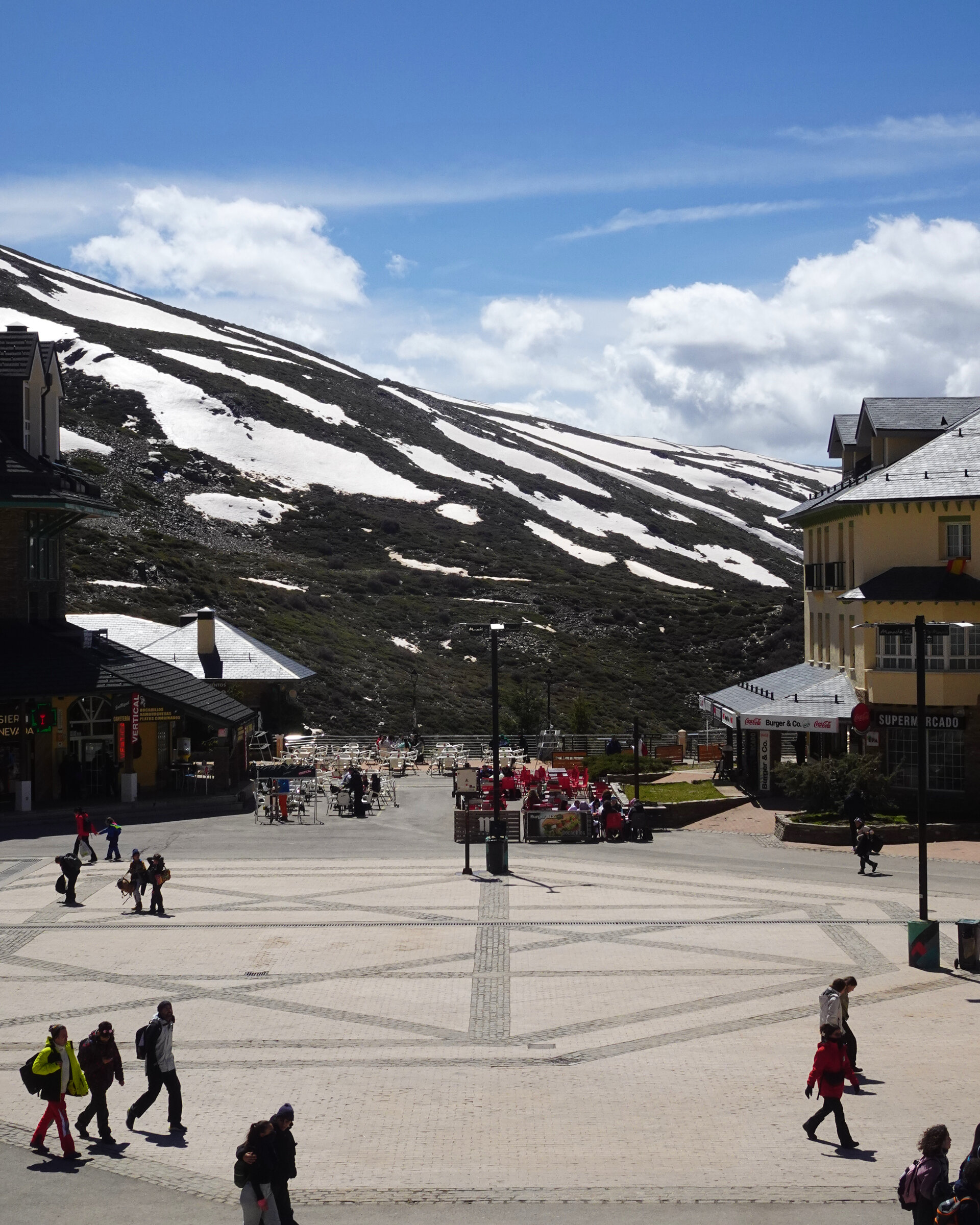 People crossing a plaza below snow-capped mountains