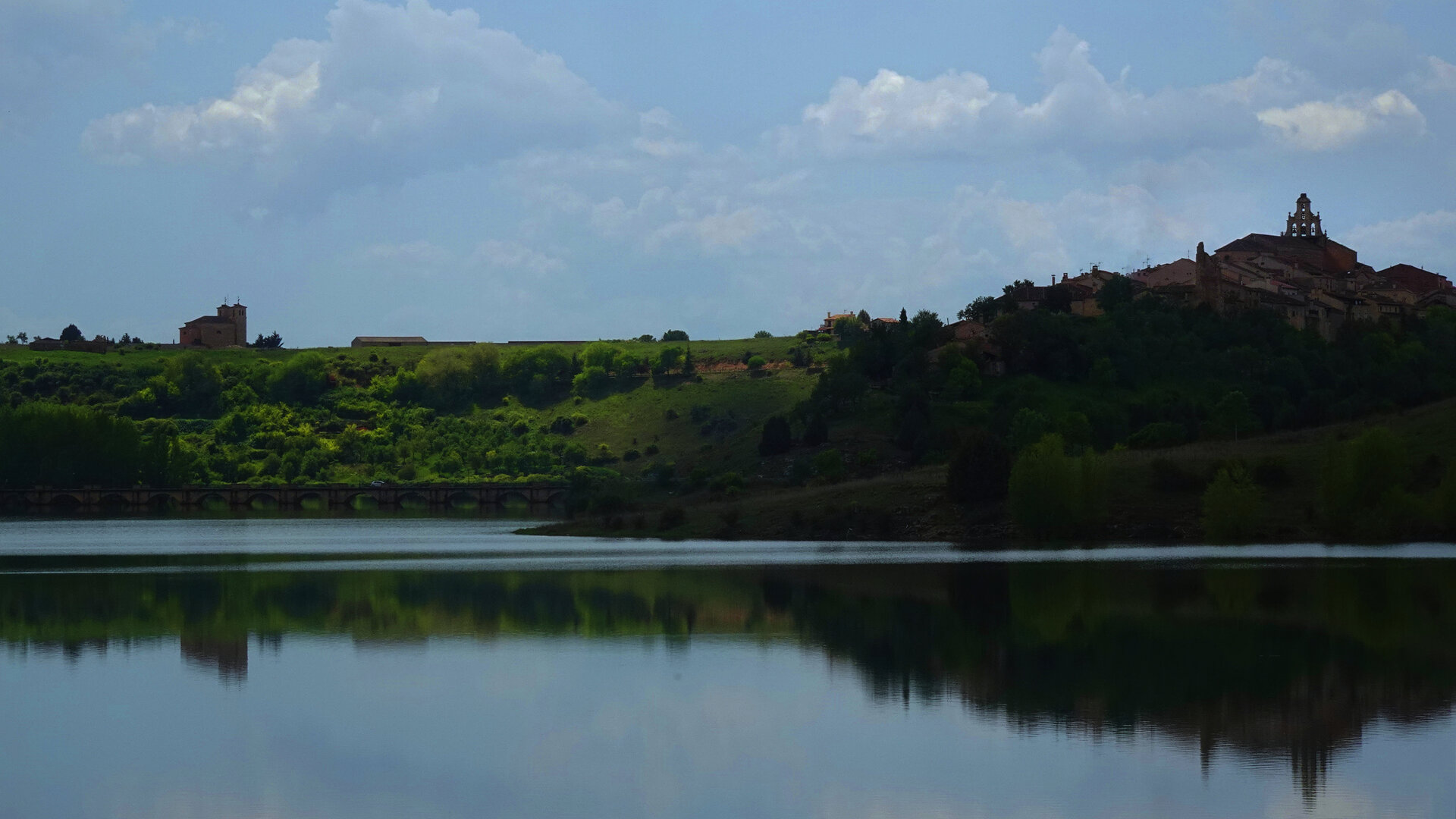 A calm lake below a wooded hill topped by a small tower