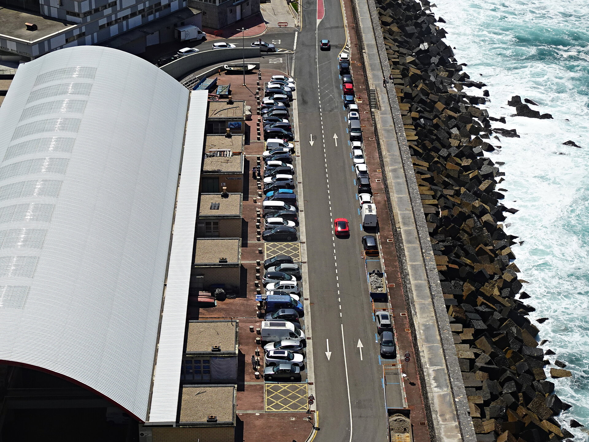 Aerial view of a seafront road and curved-roof building beside a rocky breakwater