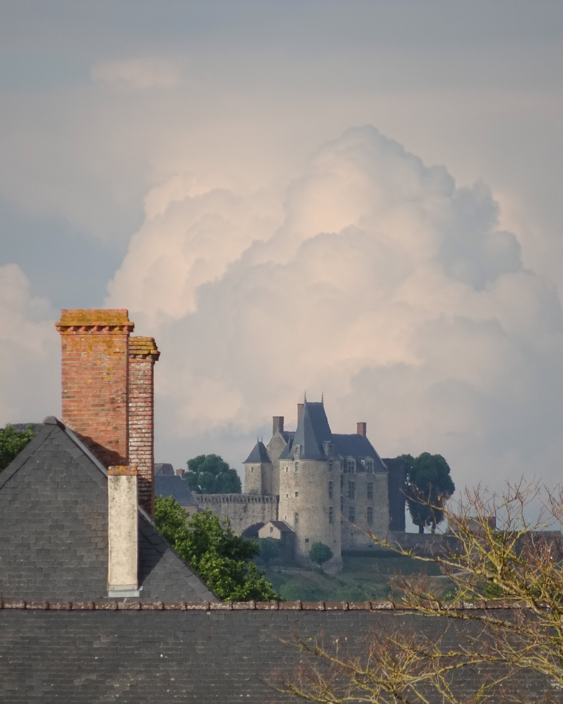 A stone château with towers rising above village rooftops and a billowing cloud
