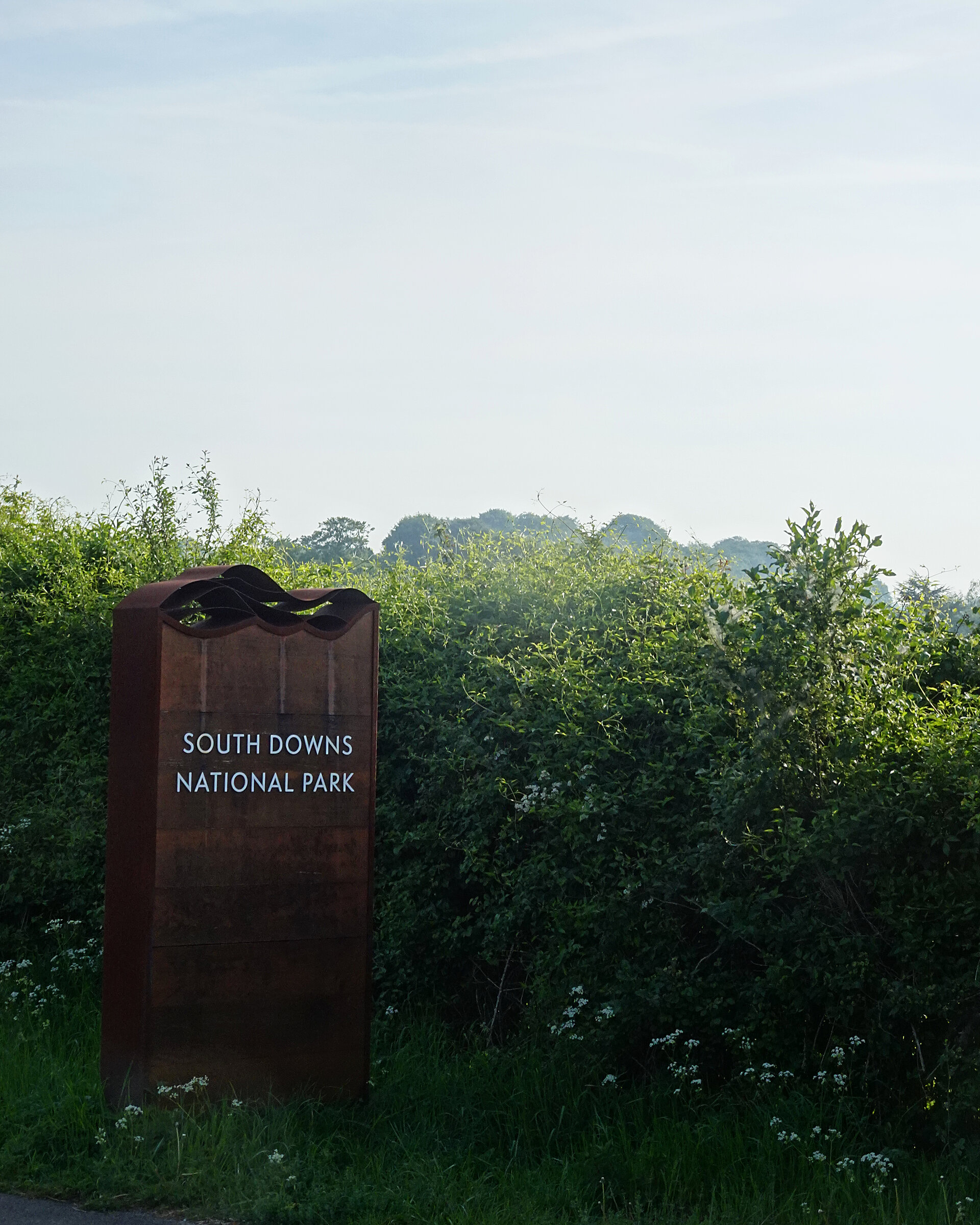 A rusted South Downs National Park sign beside a green hedgerow under a hazy sky