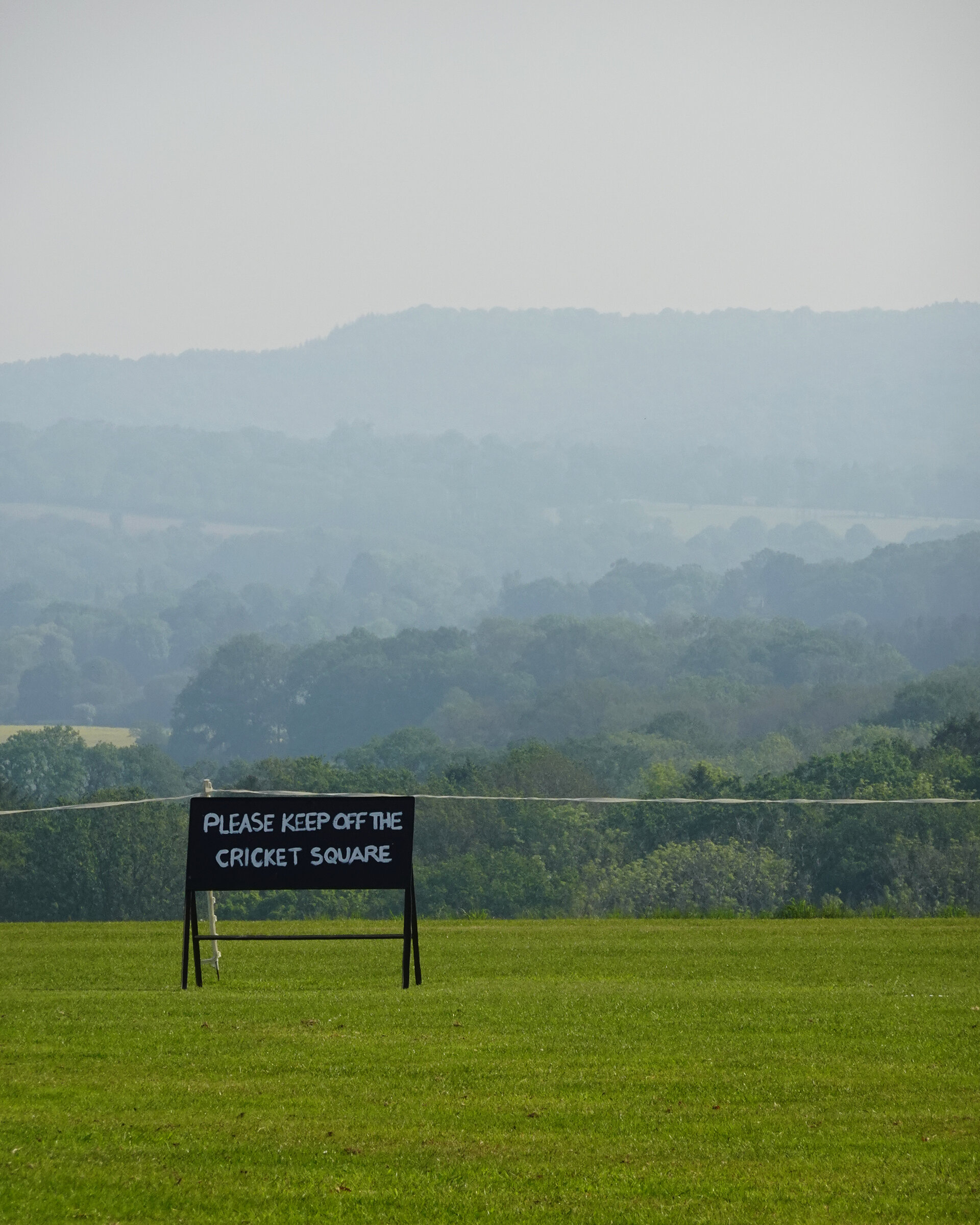 A 'keep off the cricket square' sign on a green pitch before hazy hills