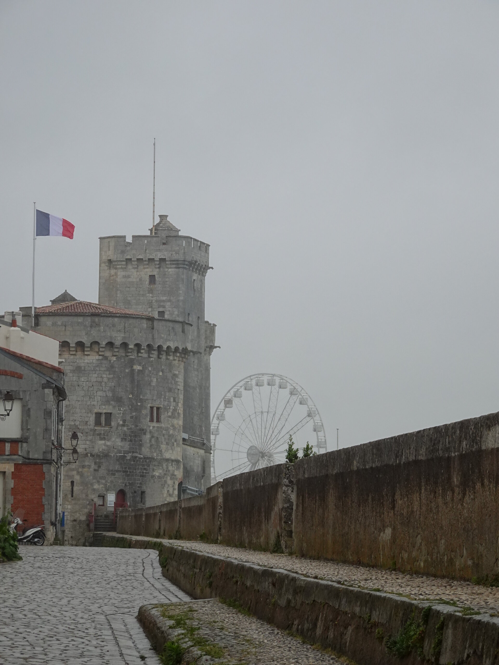 Medieval stone harbour tower flying the French flag, with a misty Ferris wheel beyond