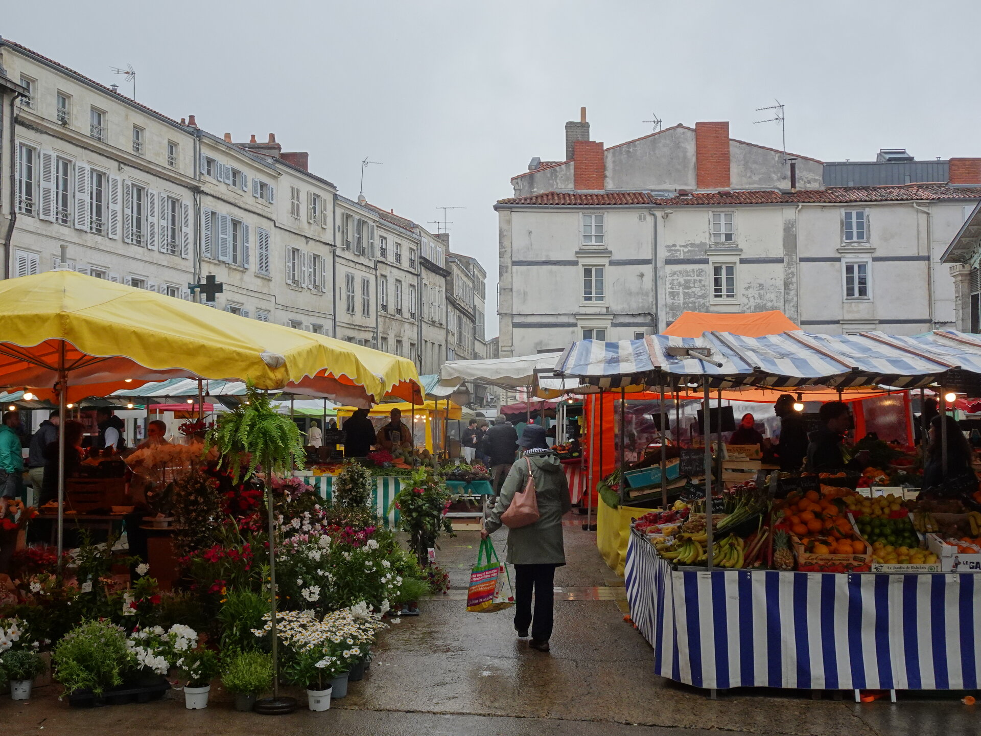 Covered market stalls of flowers and fruit on a rainy square below old town buildings