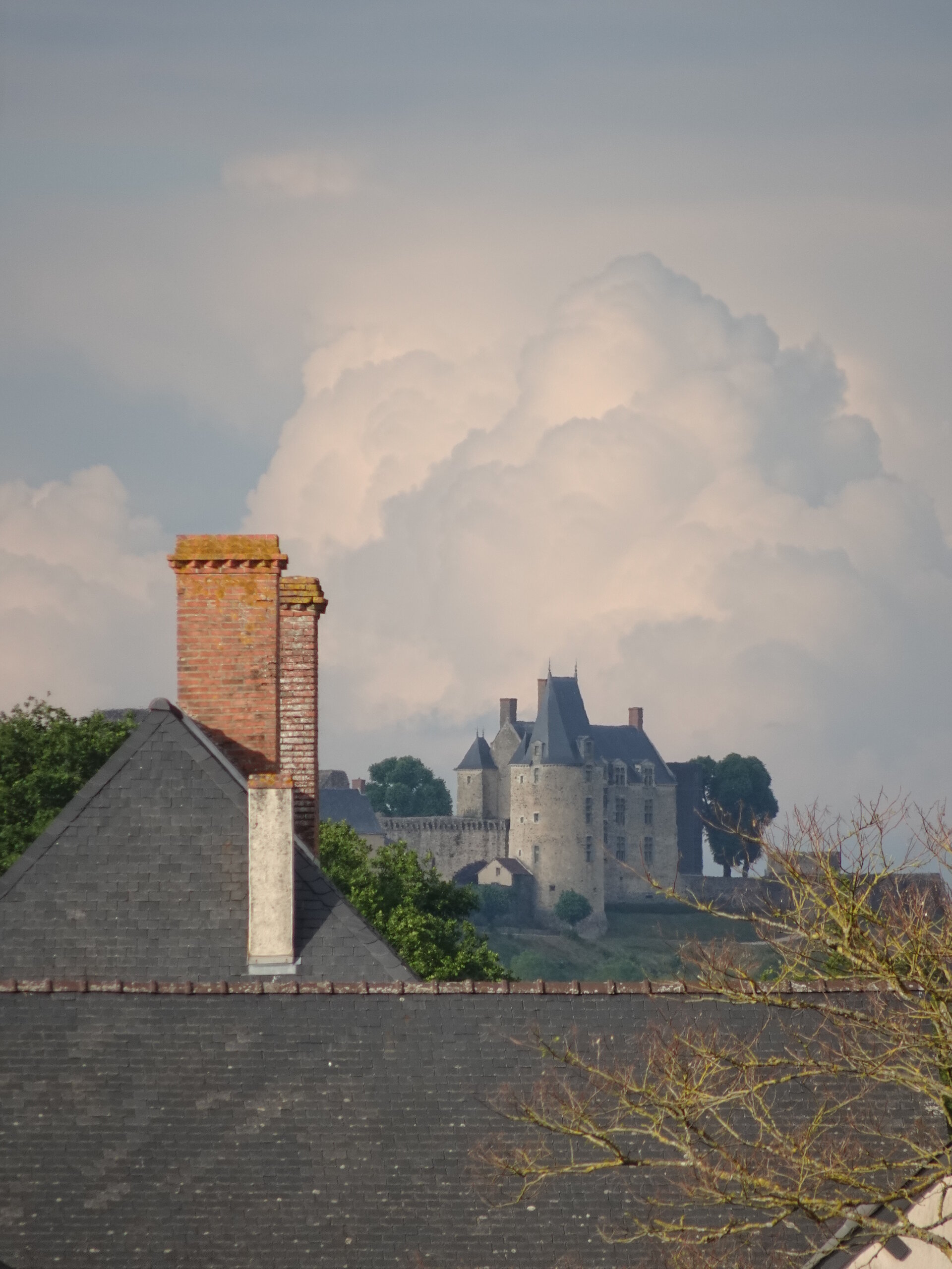 A stone château with towers rising above village rooftops and a billowing cloud