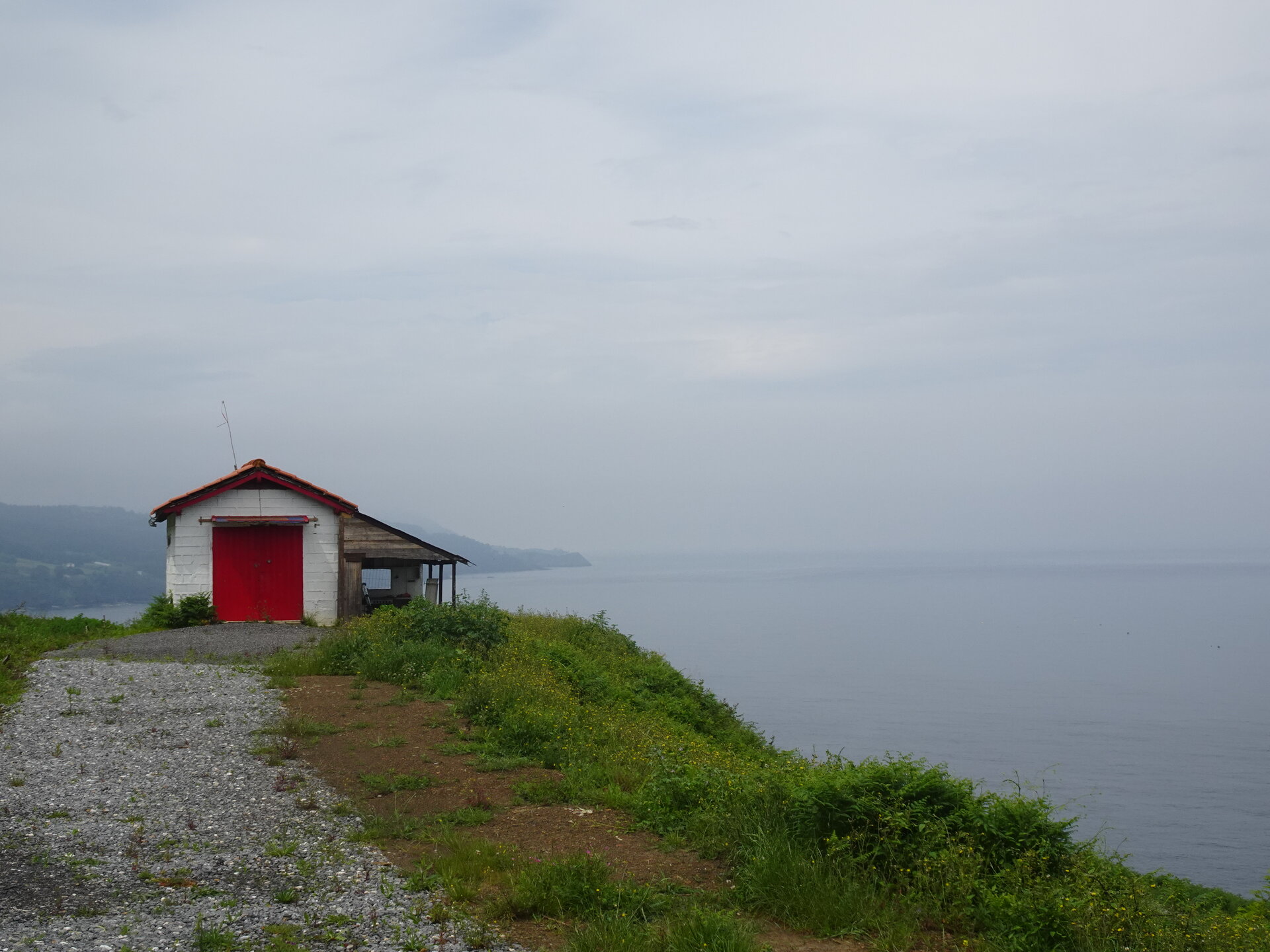White boathouse with a red door on a grassy clifftop above the sea