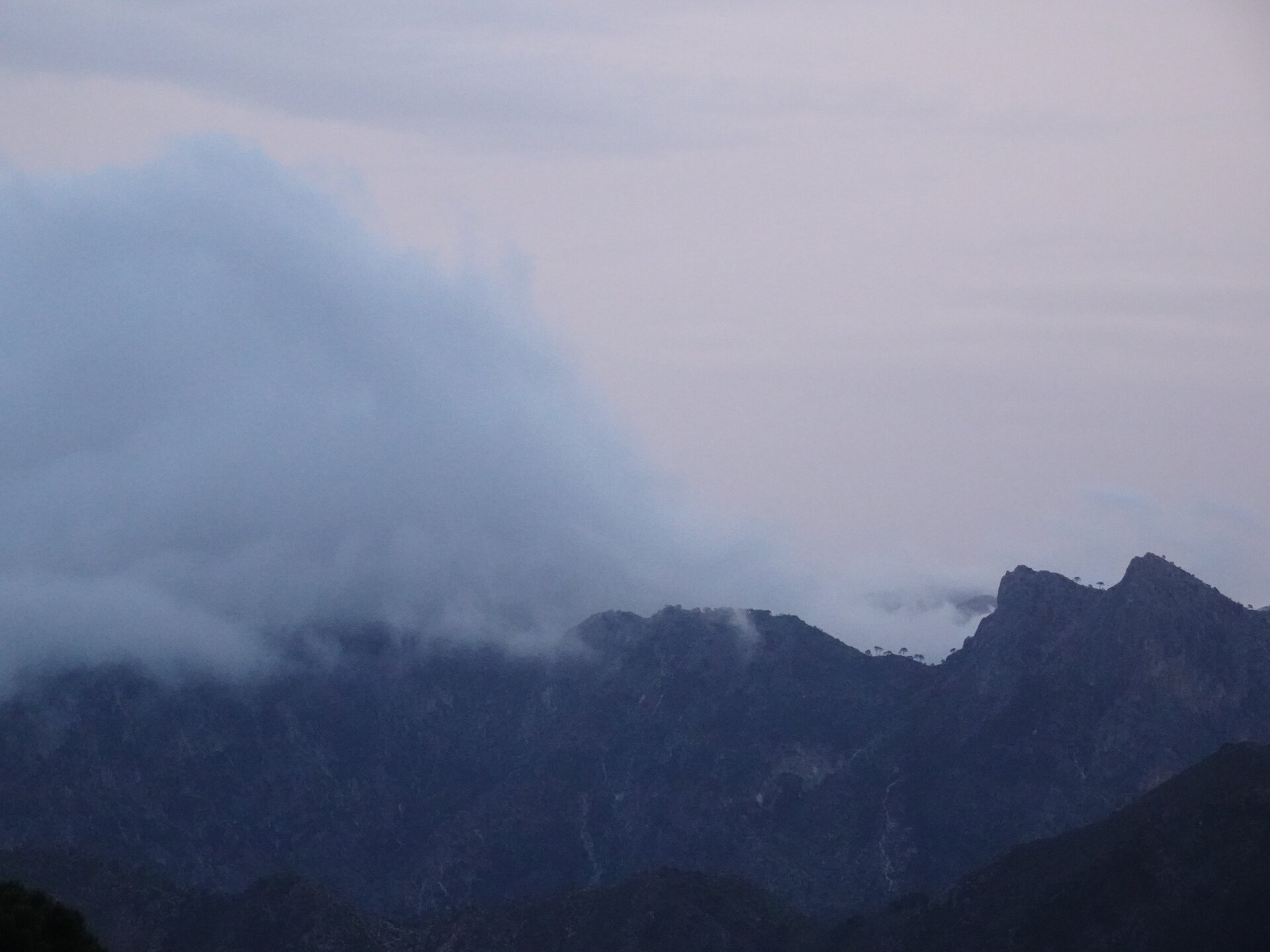 Low cloud rolling over a dark mountain ridge under a pale evening sky