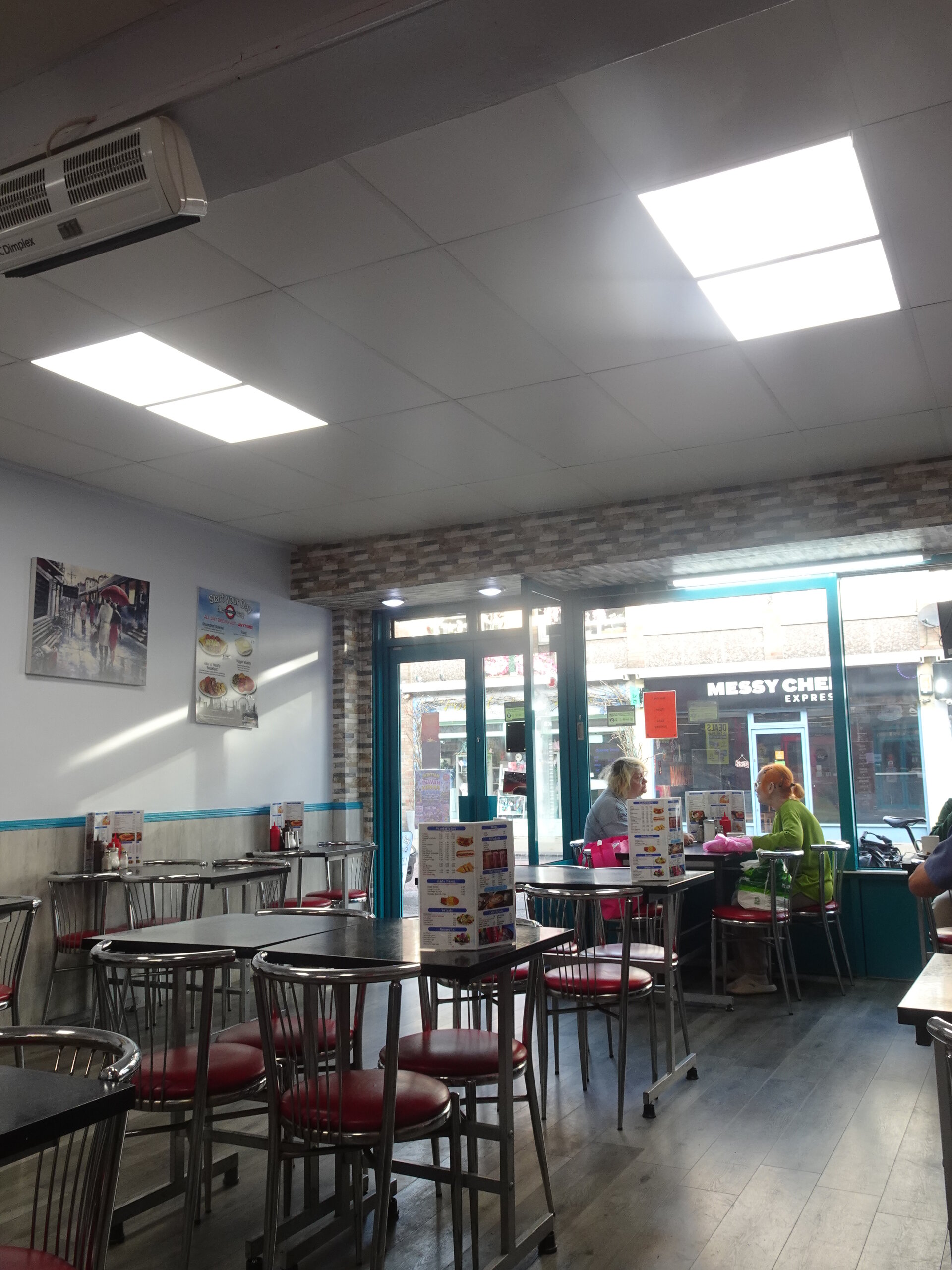 A bright greasy-spoon café interior with metal chairs, red seats and diners by the window
