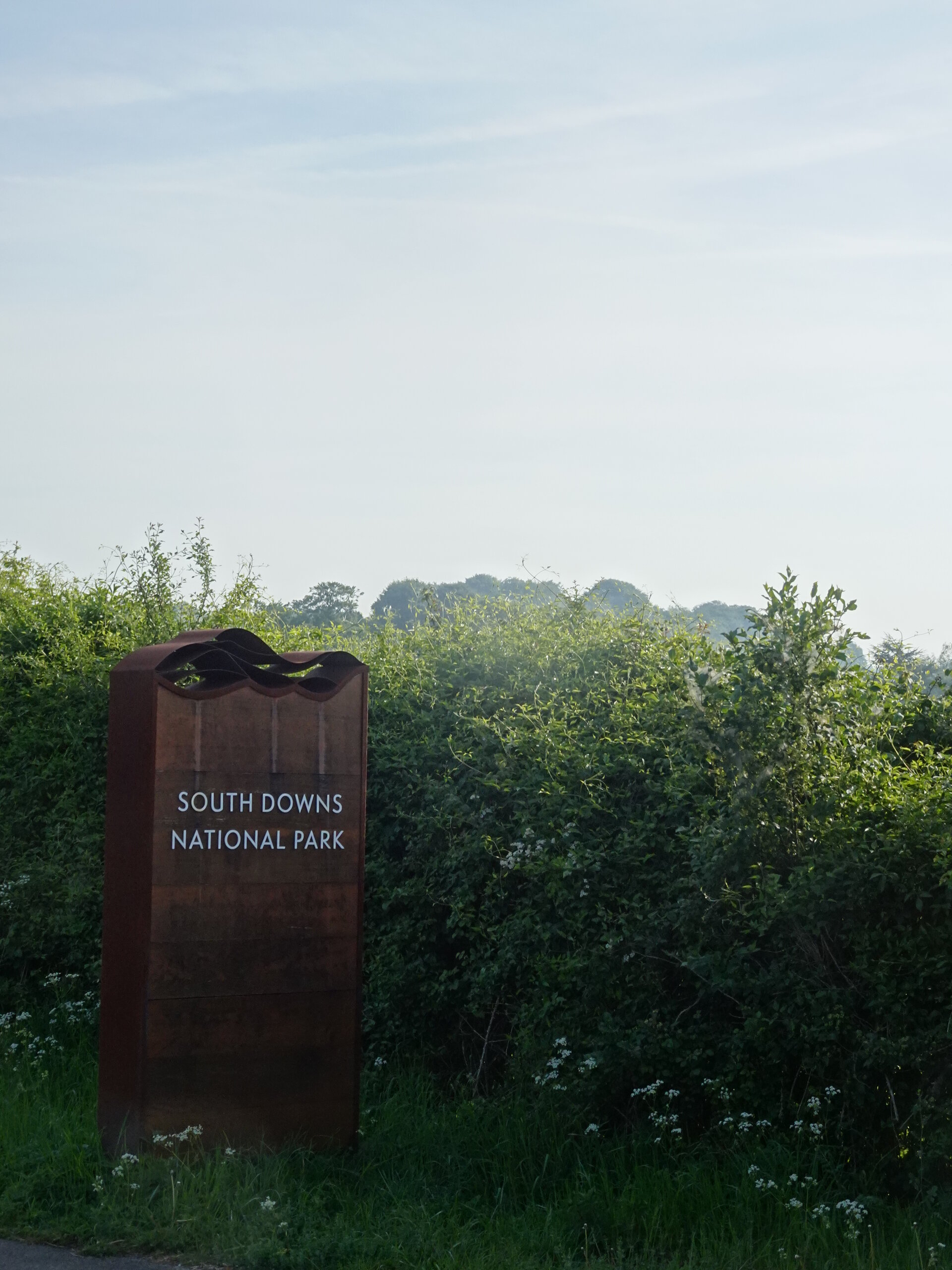 A rusted South Downs National Park sign beside a hedgerow under a hazy sky