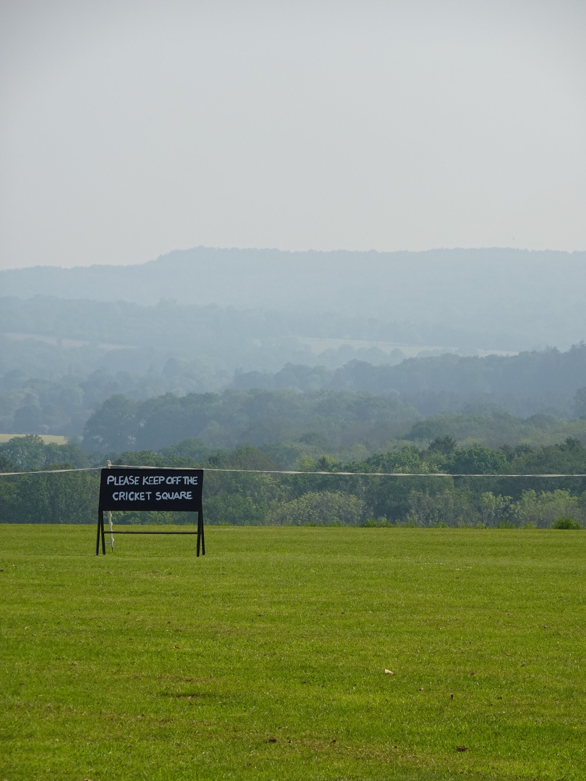 A 'keep off the cricket square' sign on a green pitch before hazy hills