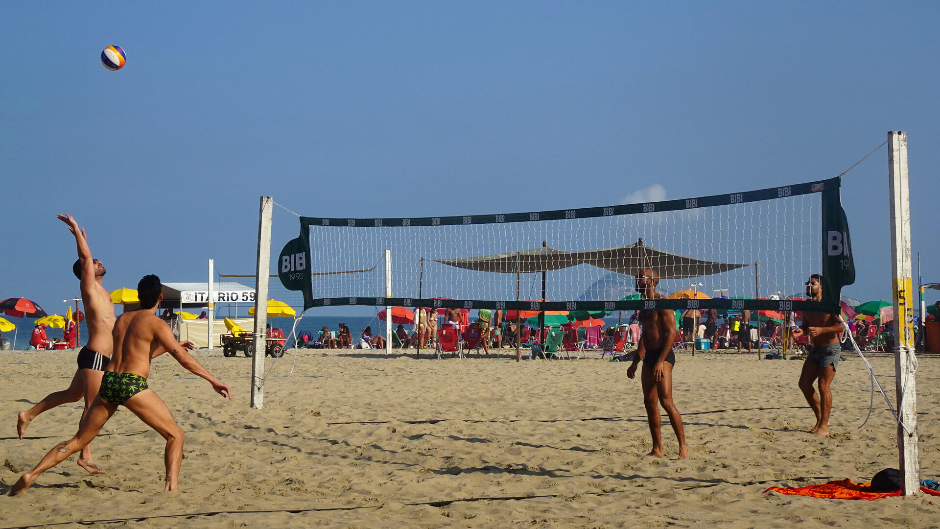 Four players either side of a beach volleyball net with parasols and sea behind