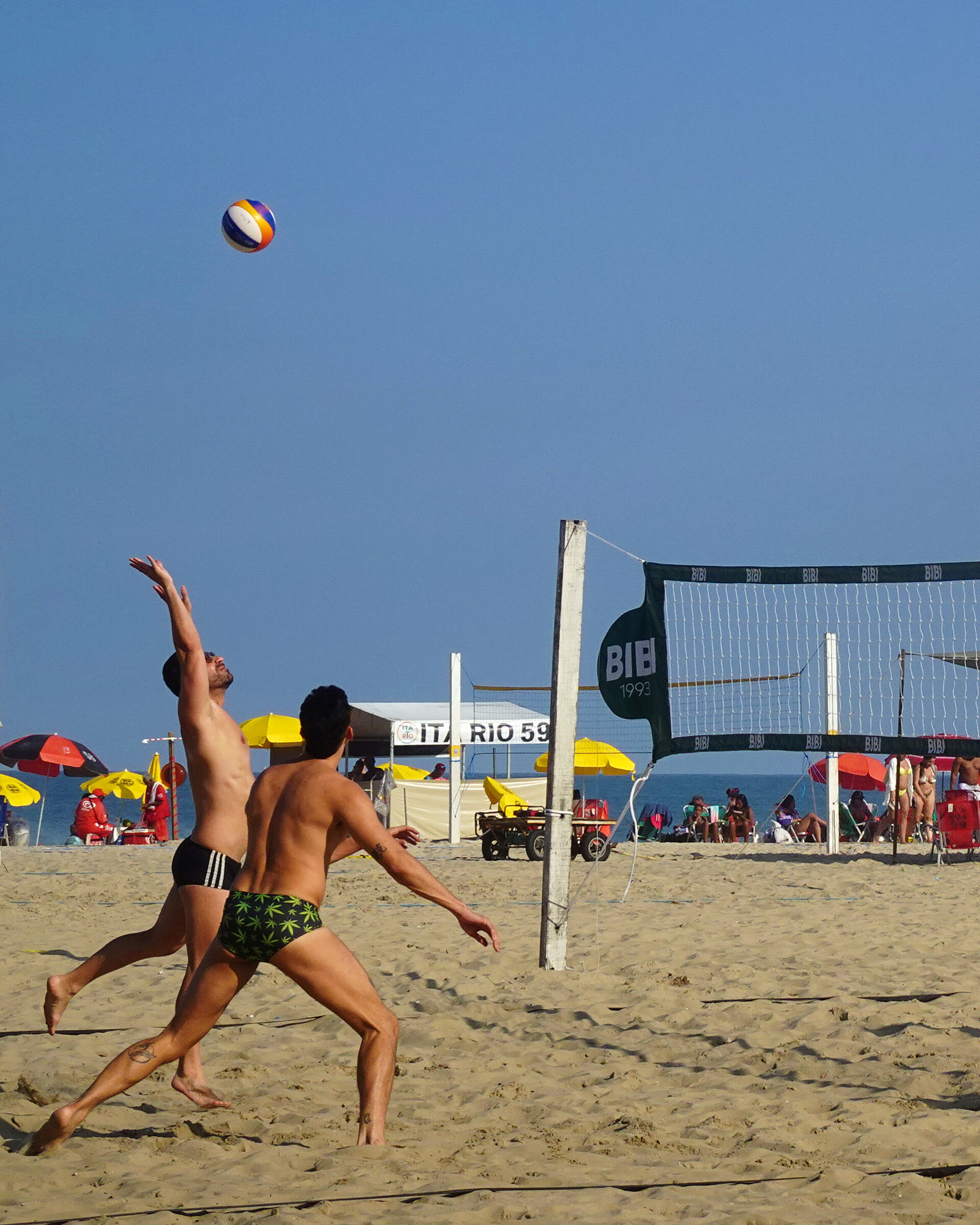 A man leaping to serve a volleyball against a clear blue sky on the beach