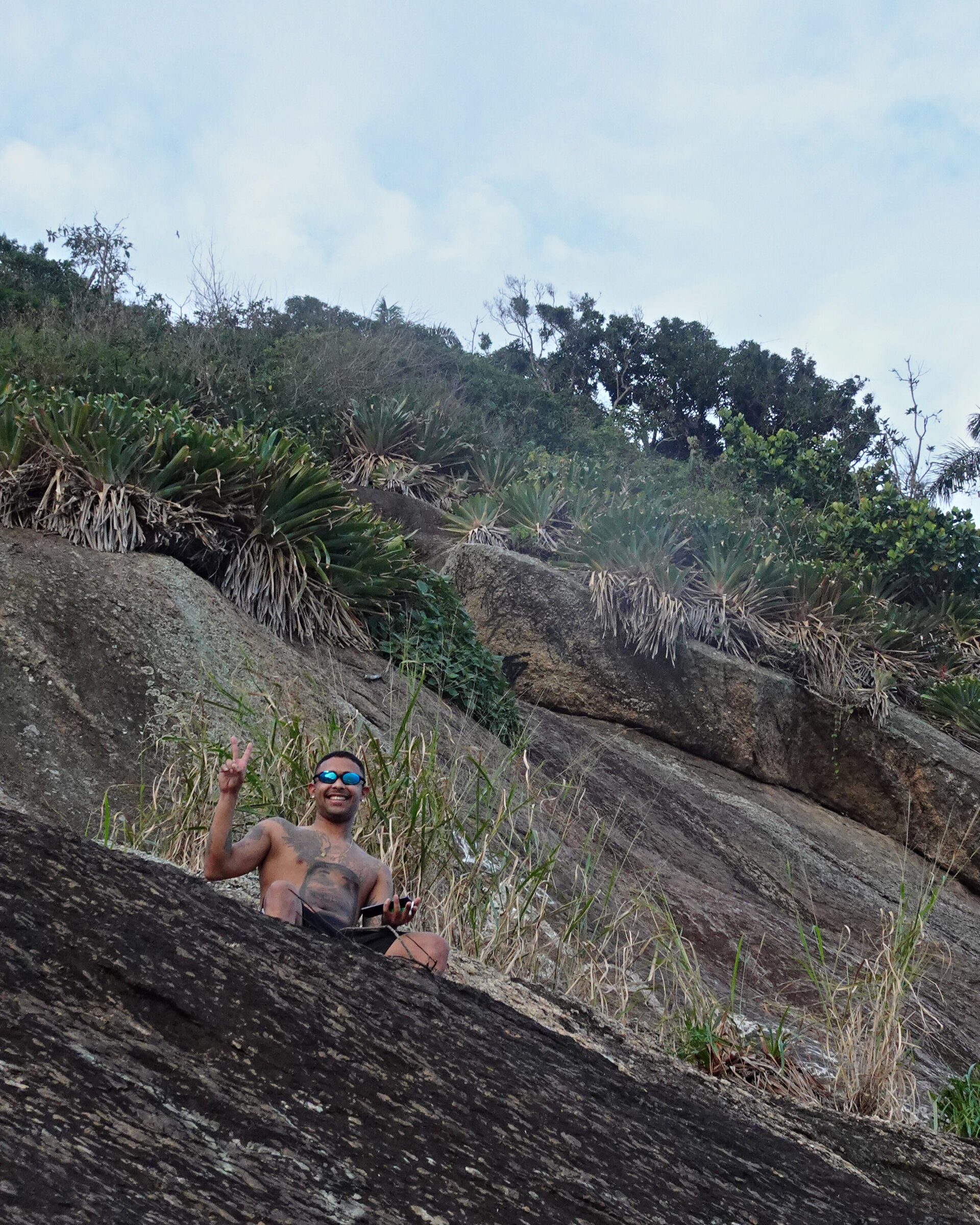 A man sitting on a bare rock slope below palms, grinning and holding up a peace sign