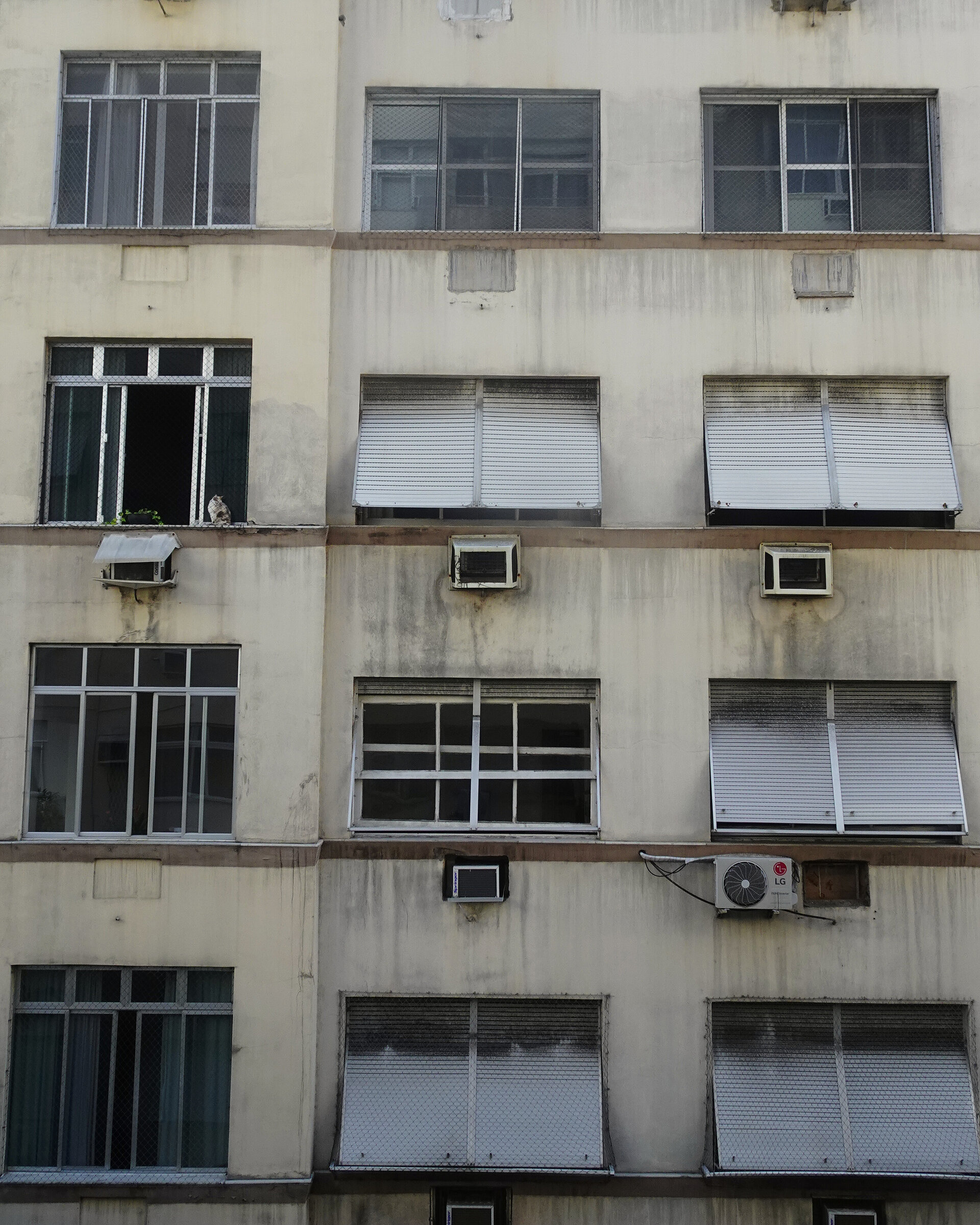 A weathered apartment block facade of shutters and air conditioning units, a cat in one window