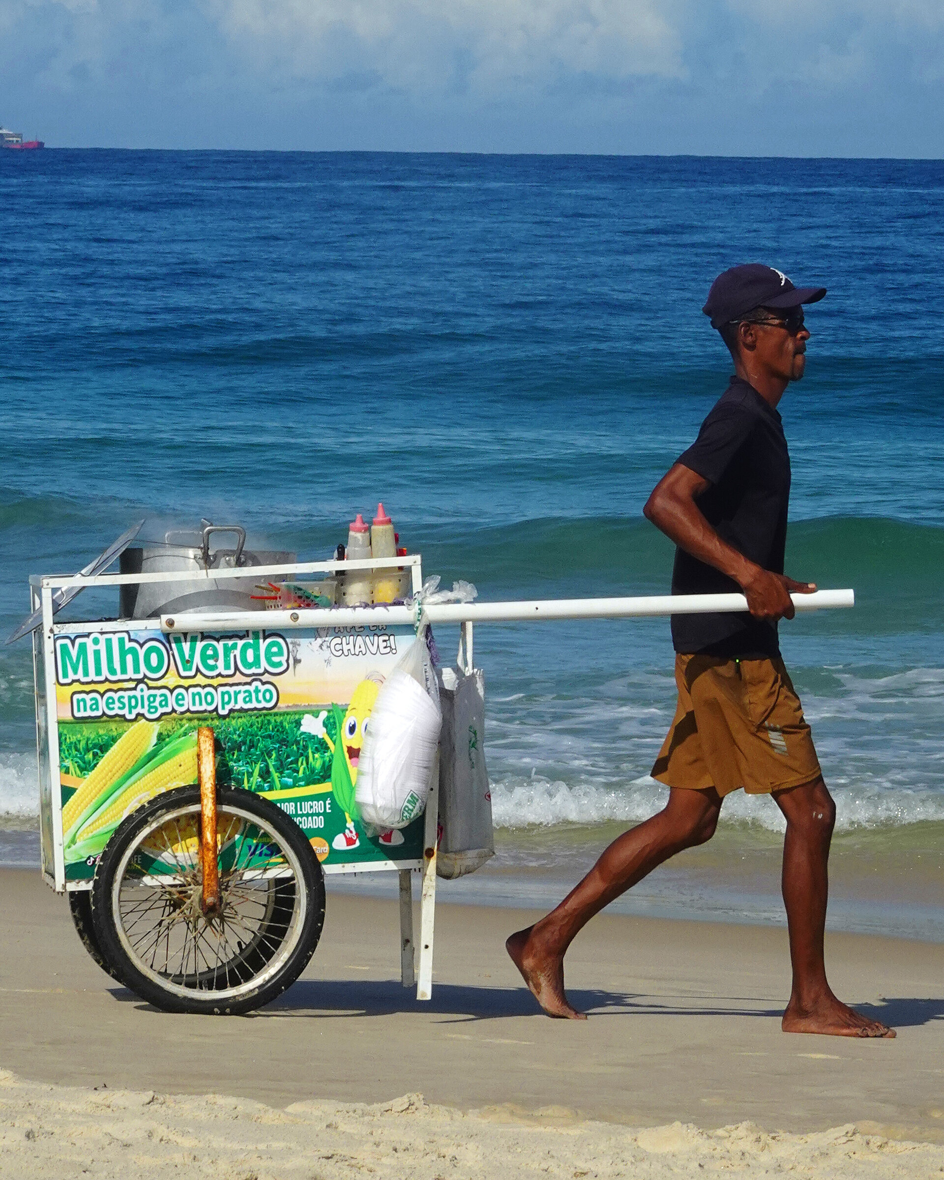 A barefoot vendor pulling a Milho Verde corn cart along the sand by the sea