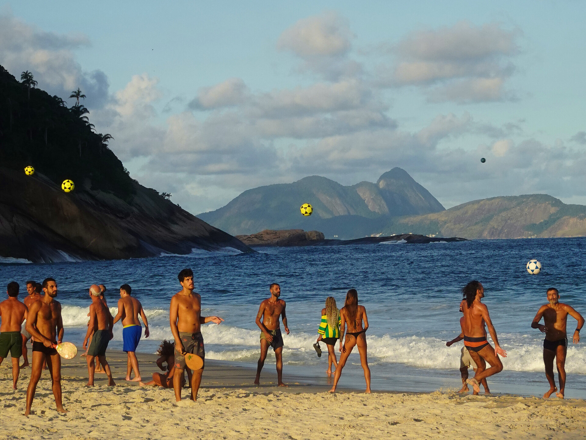 People playing footvolley and frescobol at the shoreline with mountains across the bay